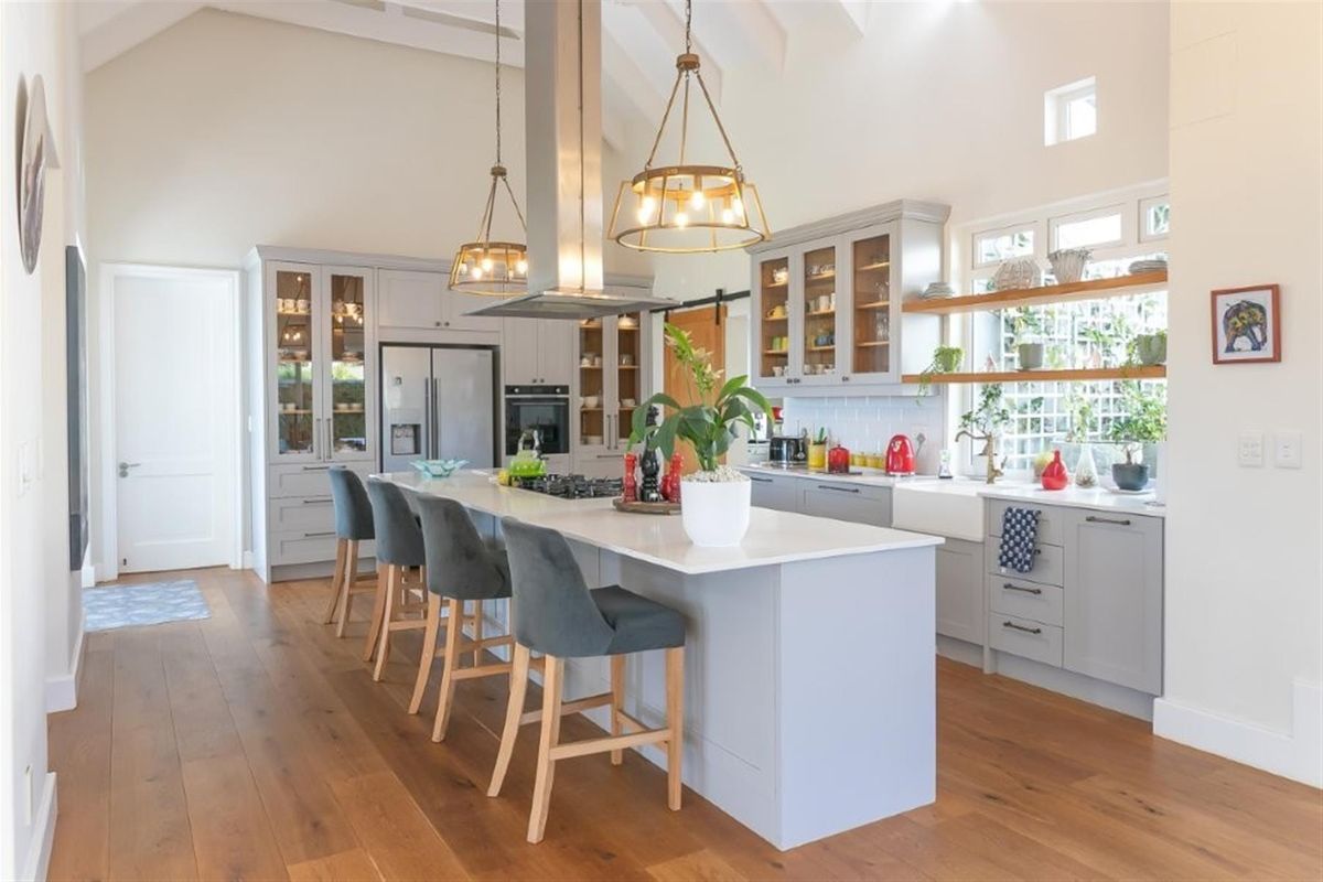 Modern farmhouse-style kitchen featuring a large central island, quartz countertops, farmhouse sink, shaker cabinetry, and high vaulted ceilings with timber flooring.