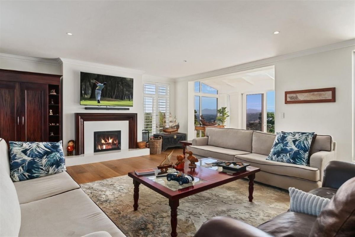 Spacious living room featuring a fireplace with a wooden mantelpiece, hardwood flooring, and large windows with plantation shutters offering scenic views.