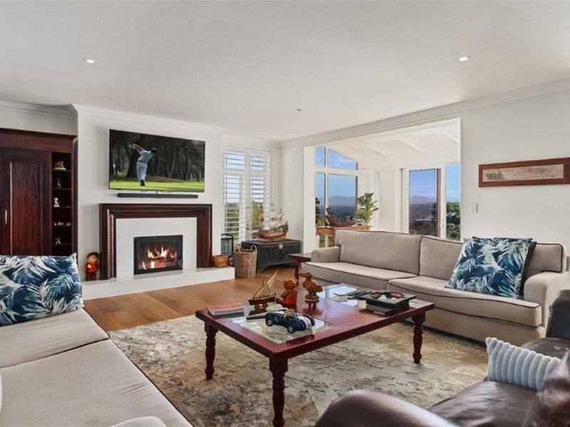 Spacious living room featuring a fireplace with a wooden mantelpiece, hardwood flooring, and large windows with plantation shutters offering scenic views.