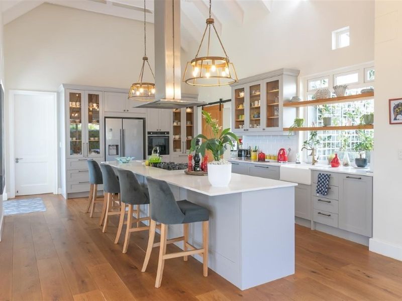 Modern farmhouse-style kitchen featuring a large central island, quartz countertops, farmhouse sink, shaker cabinetry, and high vaulted ceilings with timber flooring.