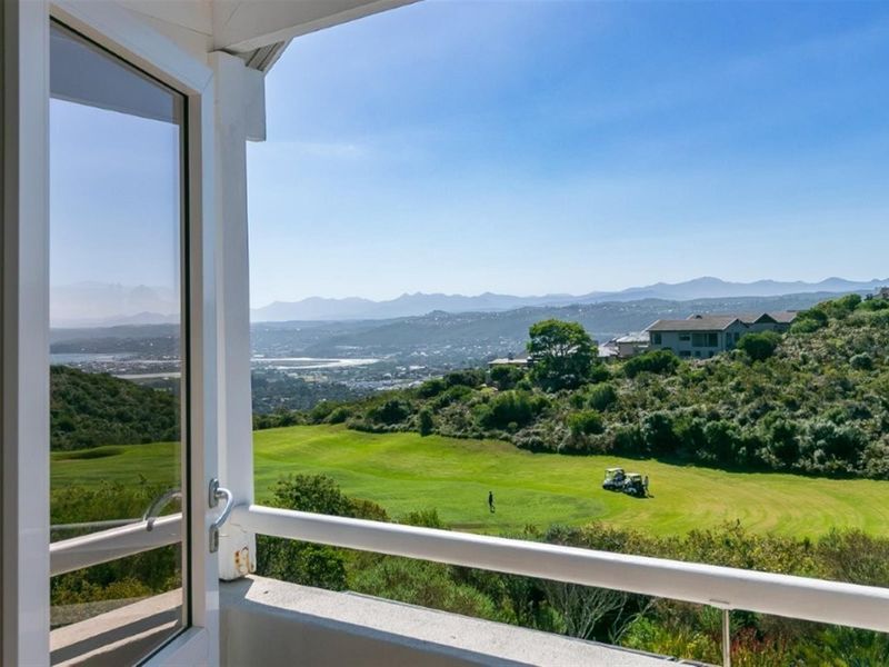 A scenic elevated view overlooking a golf course and surrounding landscape, framed by a balcony railing and glass door.