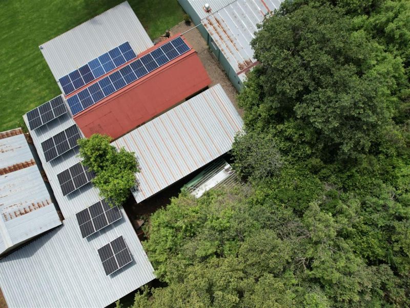 Aerial view of farm outbuildings featuring extensive solar panel installations on corrugated metal roofs, surrounded by mature trees.