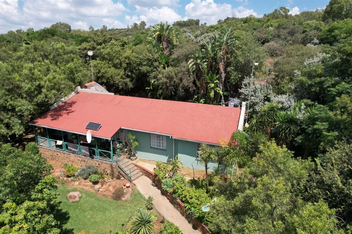 Aerial view of a farm-style dwelling featuring a red corrugated metal roof, solar panel, and a covered veranda set within a lush, wooded landscape.