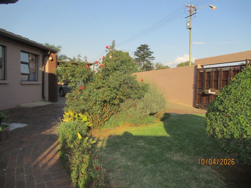 A view of the front garden area featuring a paved walkway, established landscaping, and a secure perimeter wall with a metal gate.
