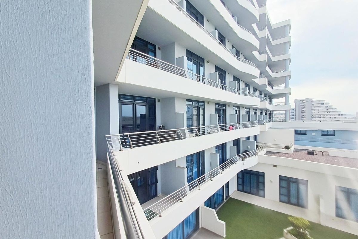 Exterior view of a modern apartment building featuring multiple levels of balconies with metal railings and a ground-level courtyard with artificial lawn.