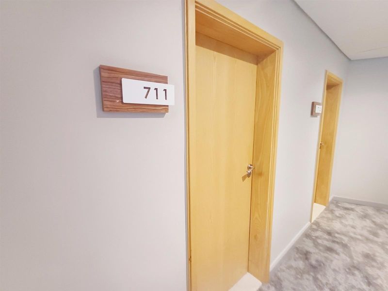 A clean, well-maintained apartment hallway featuring light wood doors, room signage, and neutral carpeting.