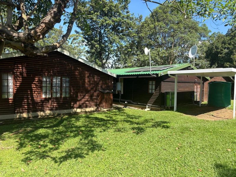 Rear view of a log cabin style property featuring a green corrugated roof, solar panels, water tank, and a carport set in a garden.