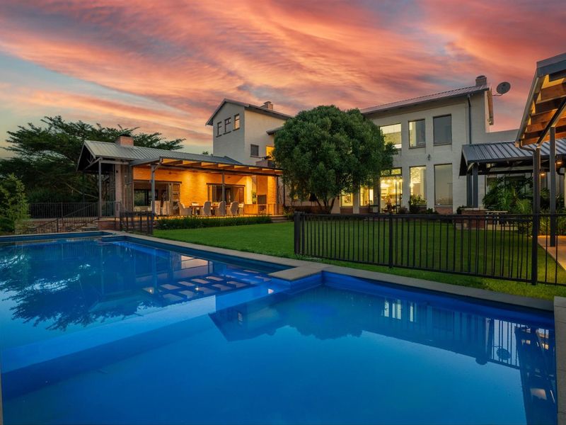 A stunning rear exterior view of the property featuring a well-maintained swimming pool, landscaped garden, and a spacious covered patio area at sunset.