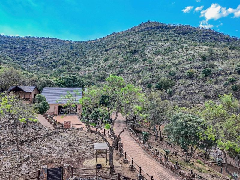 An elevated view of a small holding property featuring a paved driveway, stone-accented structures, and established trees set against a scenic mountain backdrop.