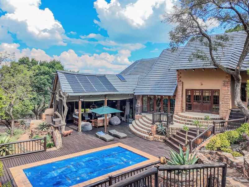 A spacious outdoor entertainment area featuring a swimming pool, extensive timber decking, covered patio space, and stone architectural details set in a natural landscape.