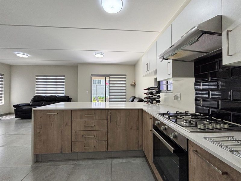 Modern open-plan kitchen featuring wood-finish cabinetry, stone countertops, gas hob, and integrated appliances, flowing seamlessly into a bright living area with tiled floors.