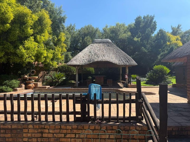 Outdoor pool area featuring a traditional thatched lapa, brick paved surround, and timber safety fencing, set within a mature garden environment.