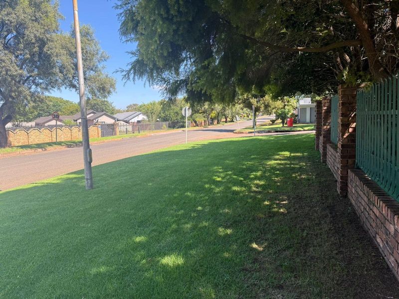 A view of the street frontage showing a well-maintained lawn, brick boundary wall with metal fencing, and surrounding neighbourhood infrastructure.
