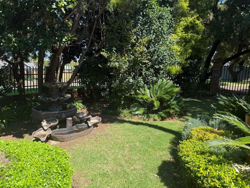 A lush, well-maintained garden featuring mature trees, landscaped shrubbery, a decorative stone water fountain, and secure wrought iron perimeter fencing.