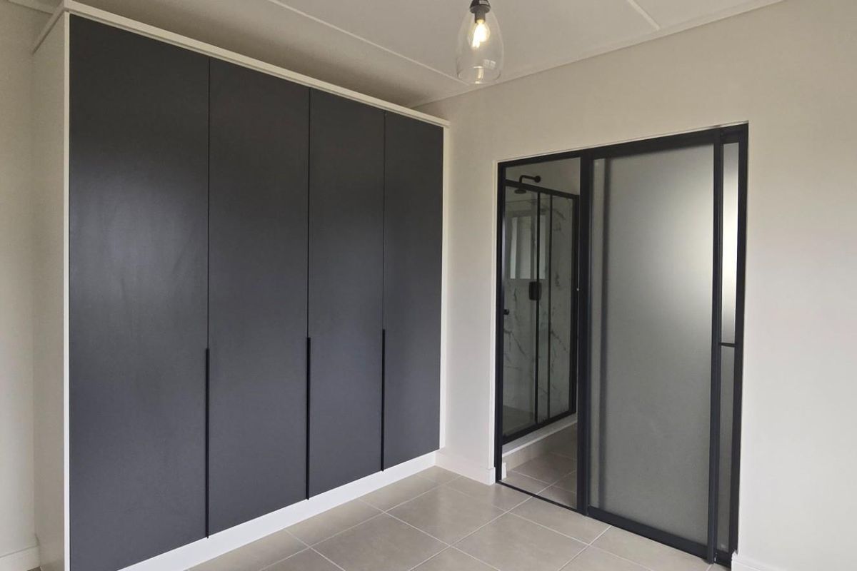 A modern bedroom featuring sleek dark built-in cupboards, tiled flooring, and a contemporary frosted glass sliding door leading to an en-suite bathroom.