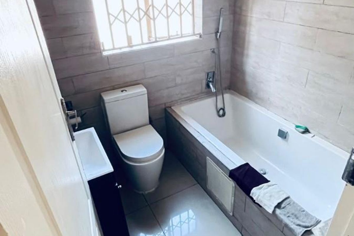 A compact bathroom featuring a built-in bathtub, a modern toilet, a vanity basin, and tiled walls with a security-grilled window.