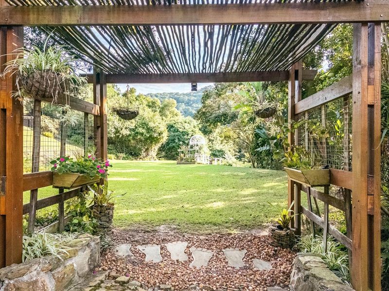 A charming wooden pergola structure framing a view of a lush, landscaped garden with a lawn and distant mountain scenery.