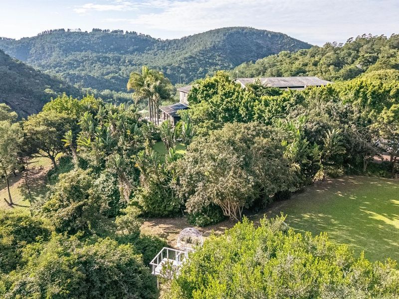 An elevated aerial view of the property showcasing the lush, landscaped garden, mature trees, and the surrounding mountainous landscape.