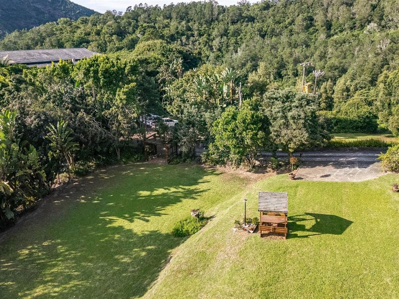 An elevated aerial view of a spacious, well-maintained garden featuring a large lawn, mature trees, and a small wooden gazebo structure.