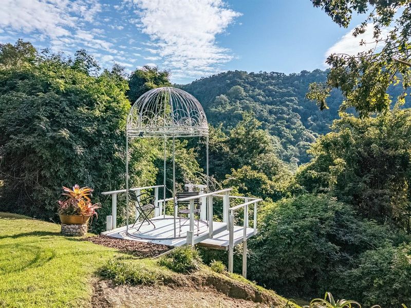 An elevated outdoor viewing platform featuring a decorative metal gazebo structure, set within a lush garden overlooking a scenic mountain landscape.