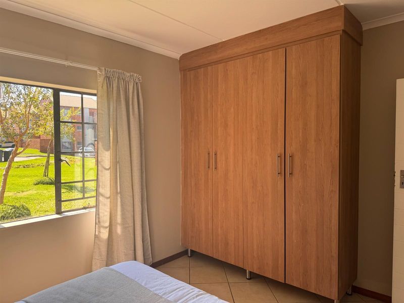 A well-lit bedroom featuring a large built-in wooden wardrobe, tiled flooring, and a window overlooking the garden area.