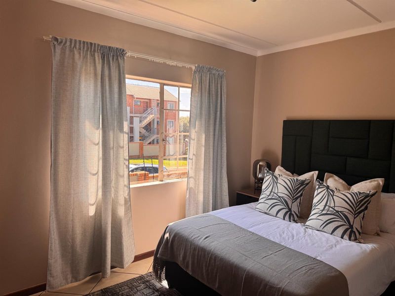 A well-lit bedroom featuring tiled flooring and a window with security bars, offering a view of the surrounding apartment complex.
