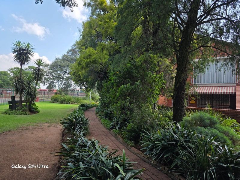A well-maintained garden featuring a brick pathway, lush green lawn, mature trees, and established shrubbery alongside the side of the brick residence.