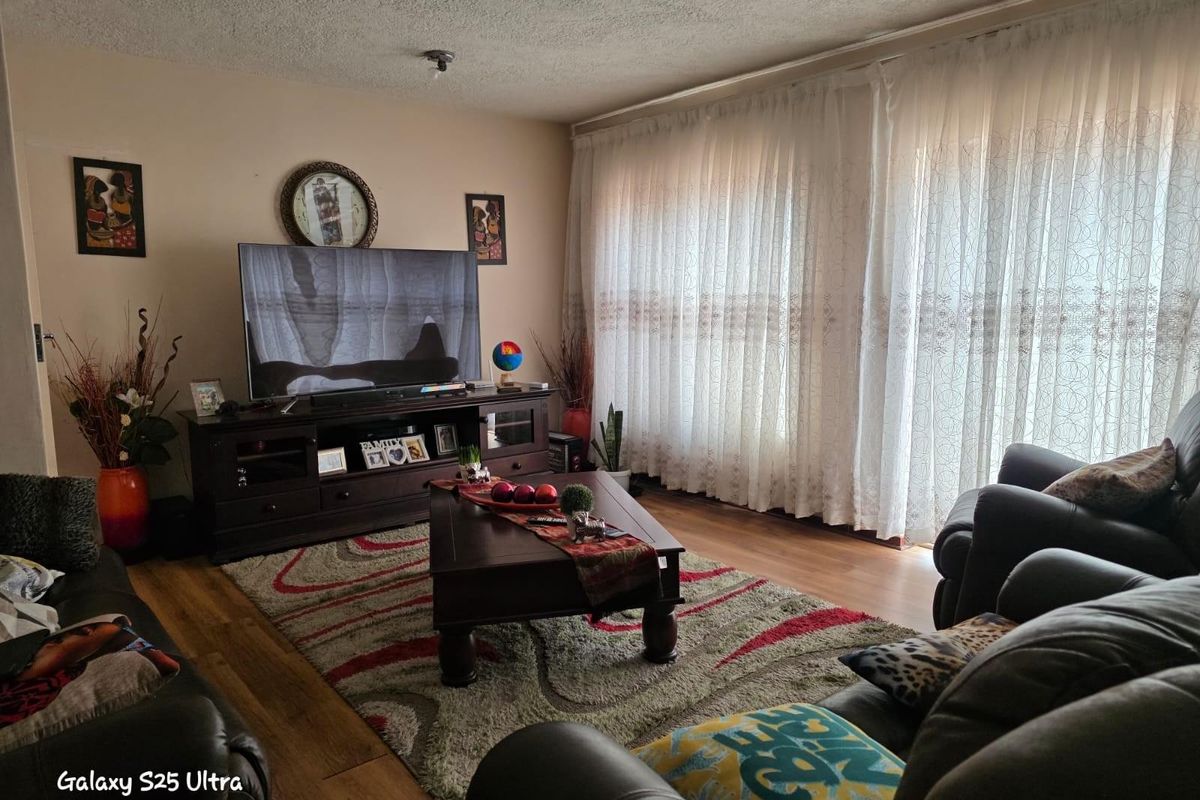 A comfortable living room featuring wood-look flooring, large windows with sheer curtains, and a neutral wall finish.