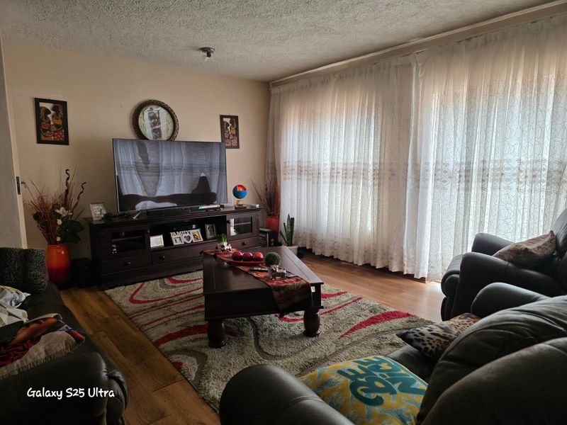 A comfortable living room featuring wood-look flooring, large windows with sheer curtains, and a neutral wall finish.