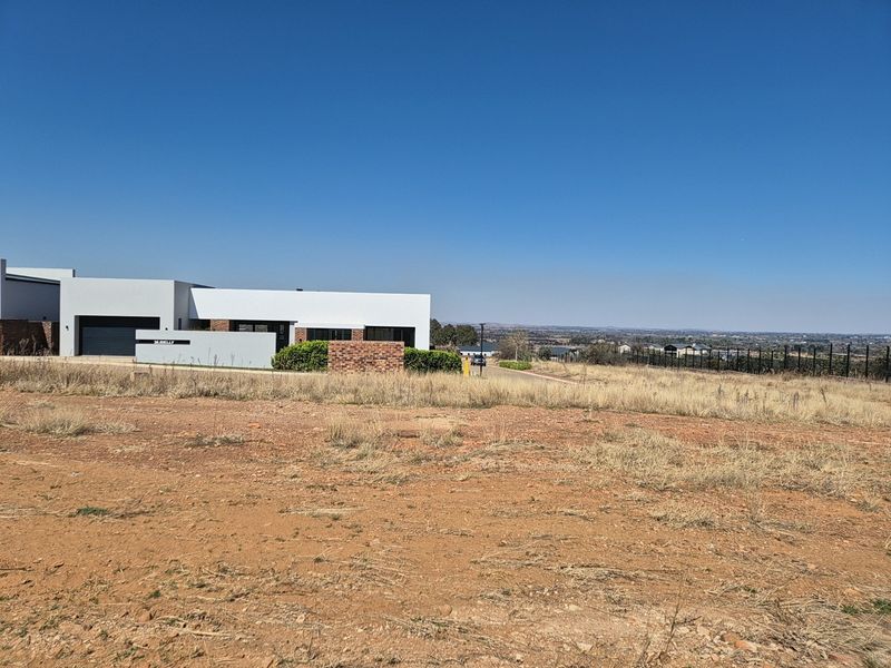 A view of a vacant residential plot featuring dry grass, perimeter fencing, and a clear view of the surrounding neighborhood and horizon.