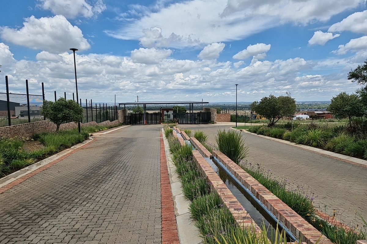 A well-maintained entrance to a residential estate featuring a paved driveway, security gate, perimeter fencing, and a decorative brick water feature.