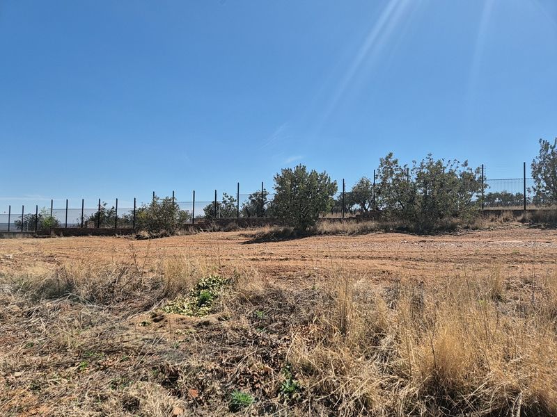 A vacant residential plot featuring natural terrain, perimeter fencing with electric security wires, and established trees along the boundary.