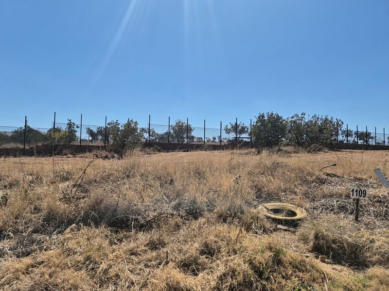 A vacant residential plot featuring dry grass, established boundary wall, and electric fencing, providing a clear space for future development.