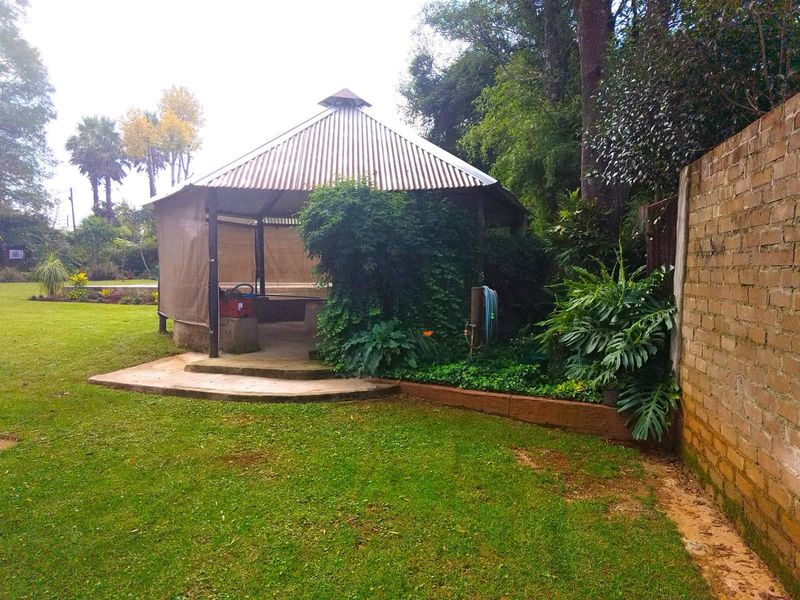 A garden gazebo with a corrugated metal roof, set within a spacious lawn area featuring mature trees and a brick boundary wall.