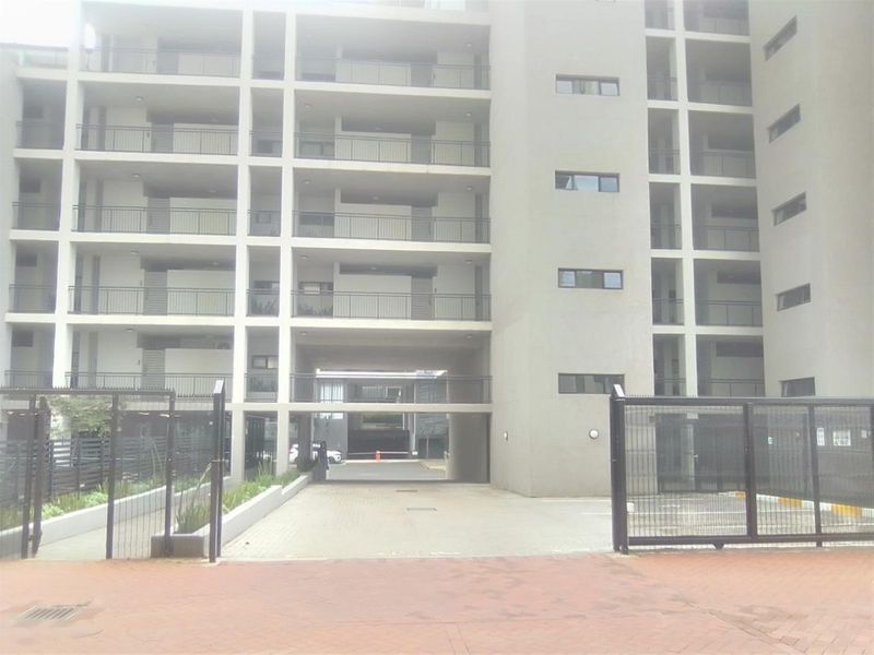 Front elevation of a modern multi-story apartment complex featuring a gated entrance, paved driveway, and multiple balconies.