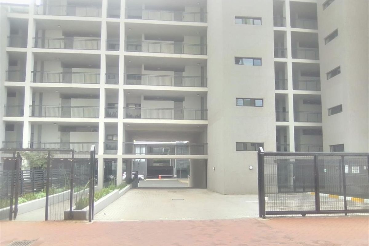 Front elevation of a modern multi-story apartment complex featuring a gated entrance, paved driveway, and multiple balconies.