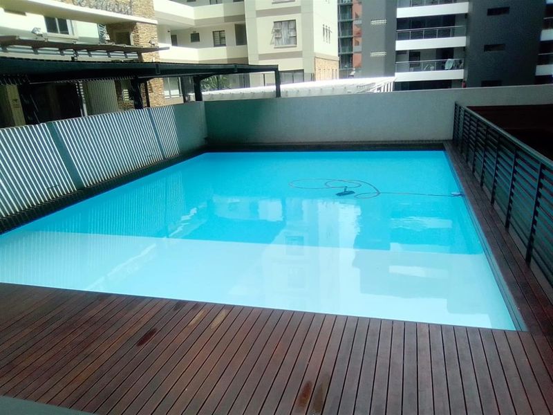 A well-maintained swimming pool area featuring timber decking and secure metal perimeter fencing, situated within an apartment complex.