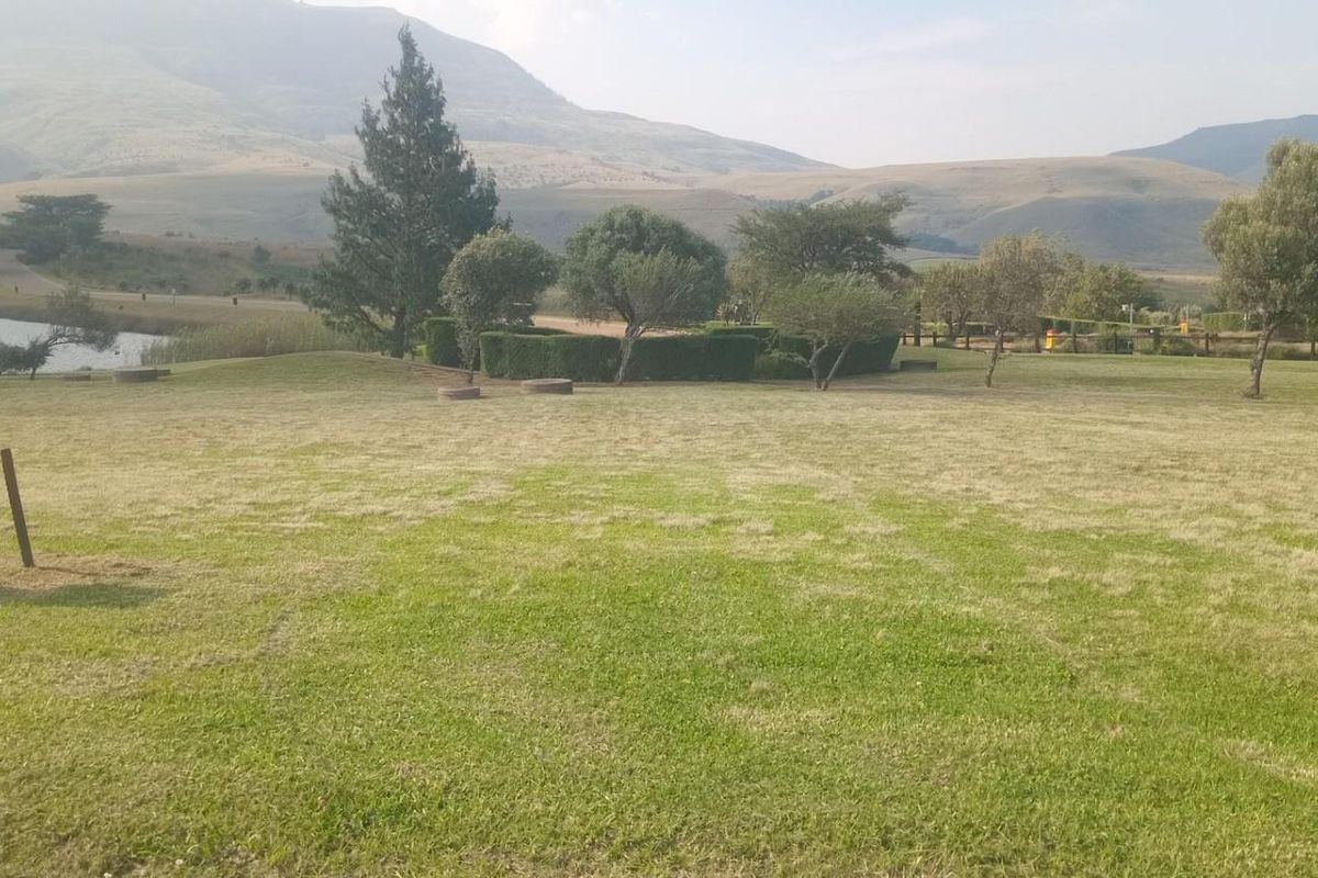 A scenic view of a vacant residential plot featuring a well-maintained lawn, mature trees, and a nearby lake with mountain vistas.
