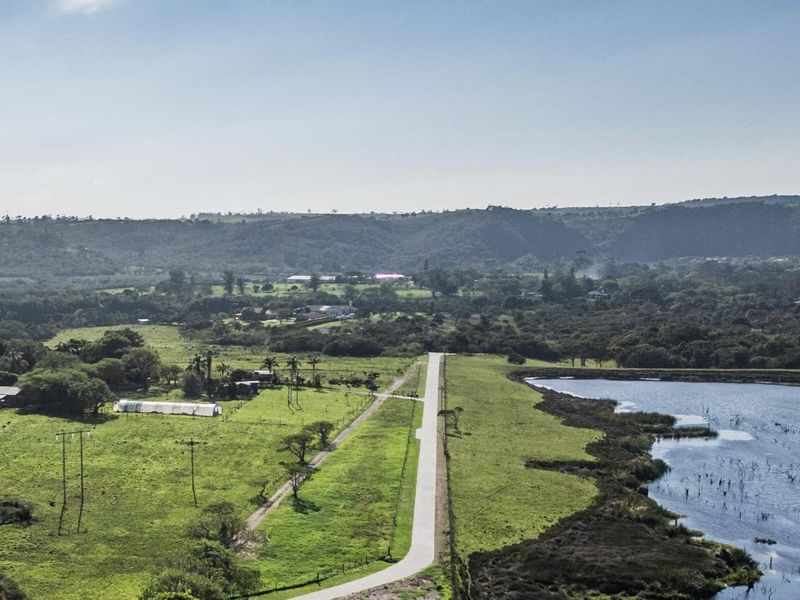 An aerial view of a vacant residential land plot featuring a paved access road, adjacent lake, and surrounding natural landscape.