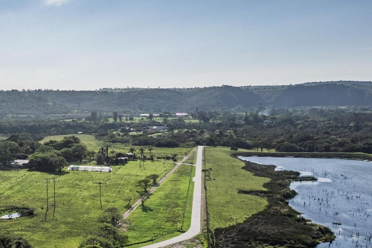 An aerial view of a vacant residential land plot featuring a paved access road, adjacent lake, and surrounding natural landscape.