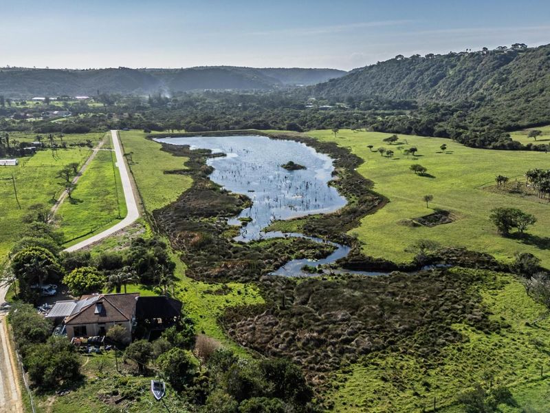 An aerial view of a large residential land parcel featuring expansive green fields, a natural lake, and surrounding hilly landscape.