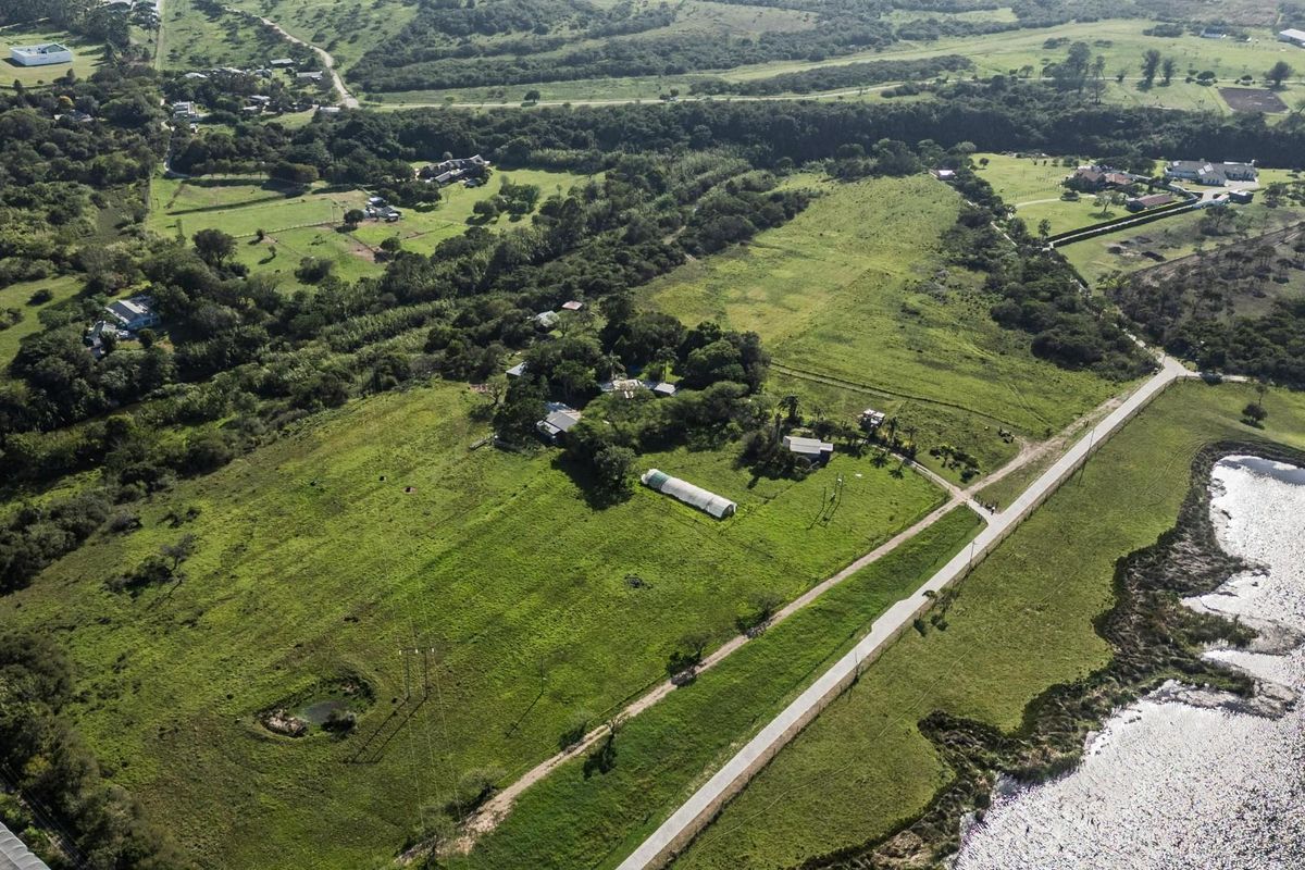 Aerial view of a large residential vacant land plot featuring open green fields, outbuildings, a greenhouse, and proximity to a water body.