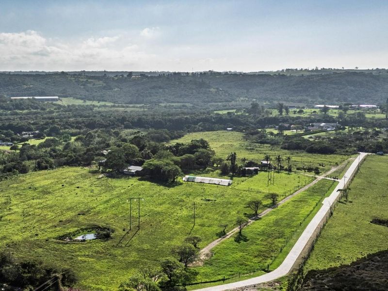 An aerial view of a large, green vacant residential plot featuring a paved road, power lines, and a small dam on the property.
