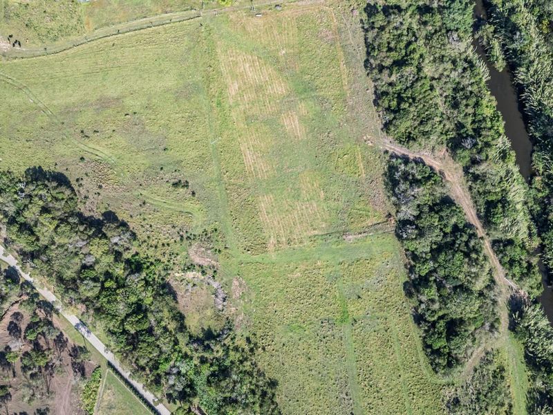 An aerial view of a vacant residential land plot featuring open grassy areas, surrounding vegetation, and proximity to a natural water course.