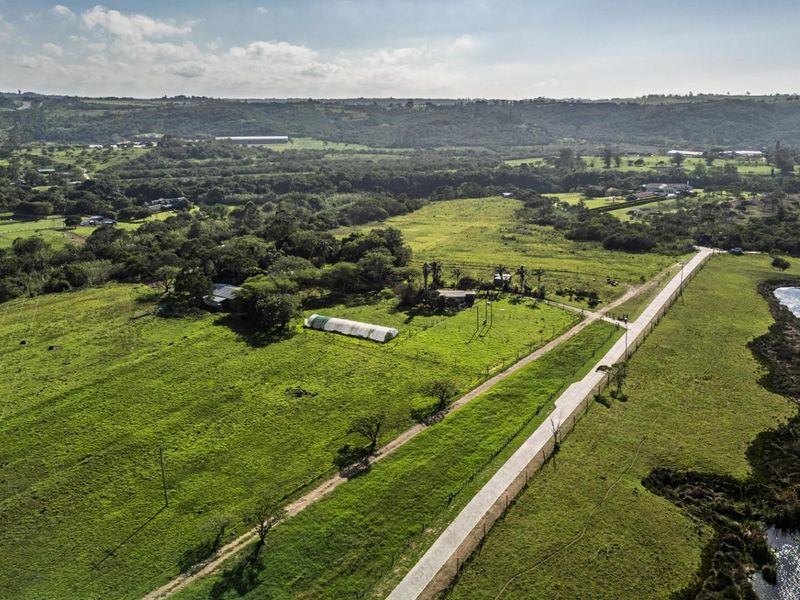 Aerial view of a large vacant residential land plot featuring open green fields, a paved access road, a small dam, and existing utility infrastructure.