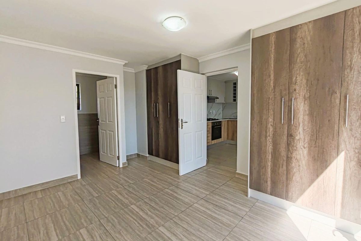 A bright bedroom featuring built-in wooden cupboards, tiled flooring, and direct access to an adjoining kitchen area.