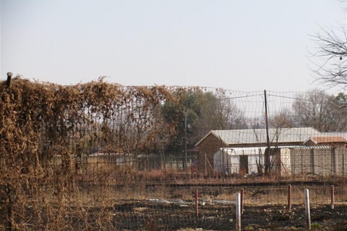 A view of a vacant residential plot featuring dry, overgrown vegetation and existing perimeter fencing with a small structure visible in the background.