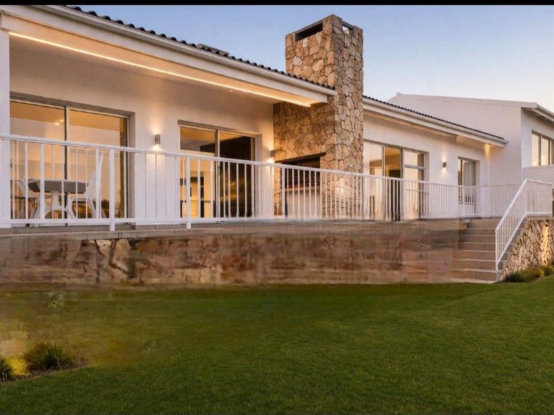 A beautifully maintained rear exterior featuring a spacious patio, stone-clad fireplace, white railings, and integrated lighting overlooking a manicured lawn.
