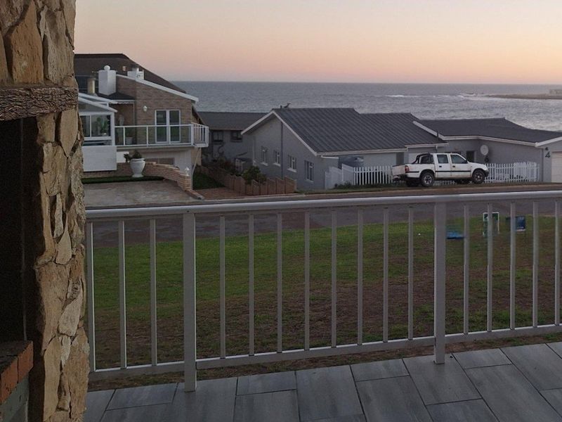 A view from a tiled patio featuring a stone-clad pillar and metal railing, overlooking a coastal neighbourhood and the ocean at sunset.