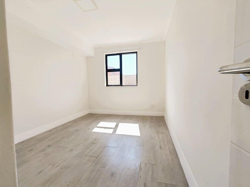 A bright, empty bedroom featuring modern light-toned wood-look flooring, neutral walls, and a window providing natural light.